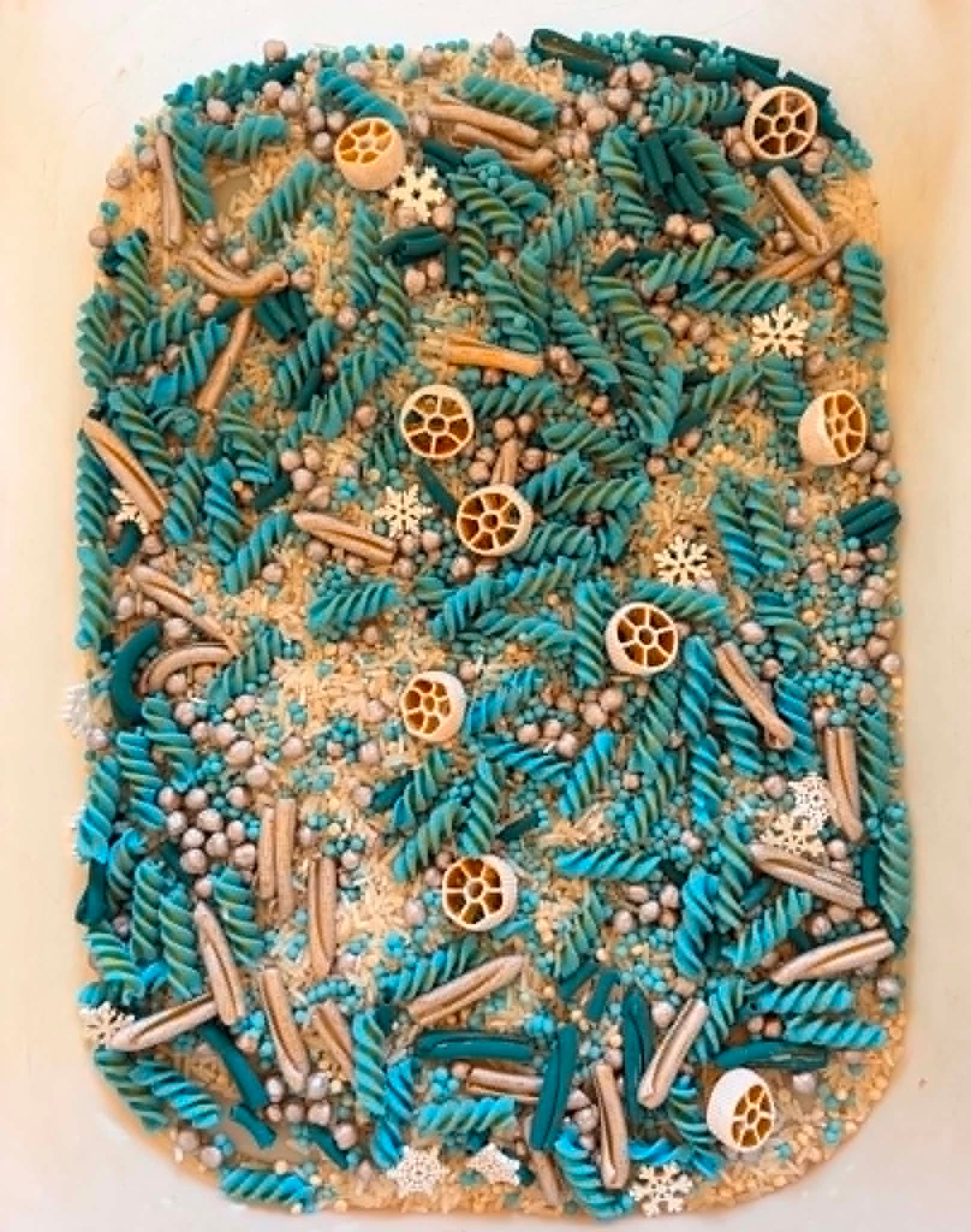 A winter sensory play tray of teal and cream dyed pasta, rice and wooden snowflakes